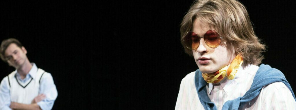 Two actors on stage; one wearing round sunglasses and a scarf, embodying Atlantic's world-class acting standard.