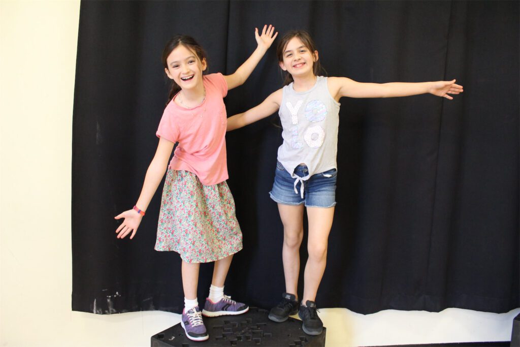 Two young girls pose with arms outstretched against a black backdrop. Sketch comedy fun!