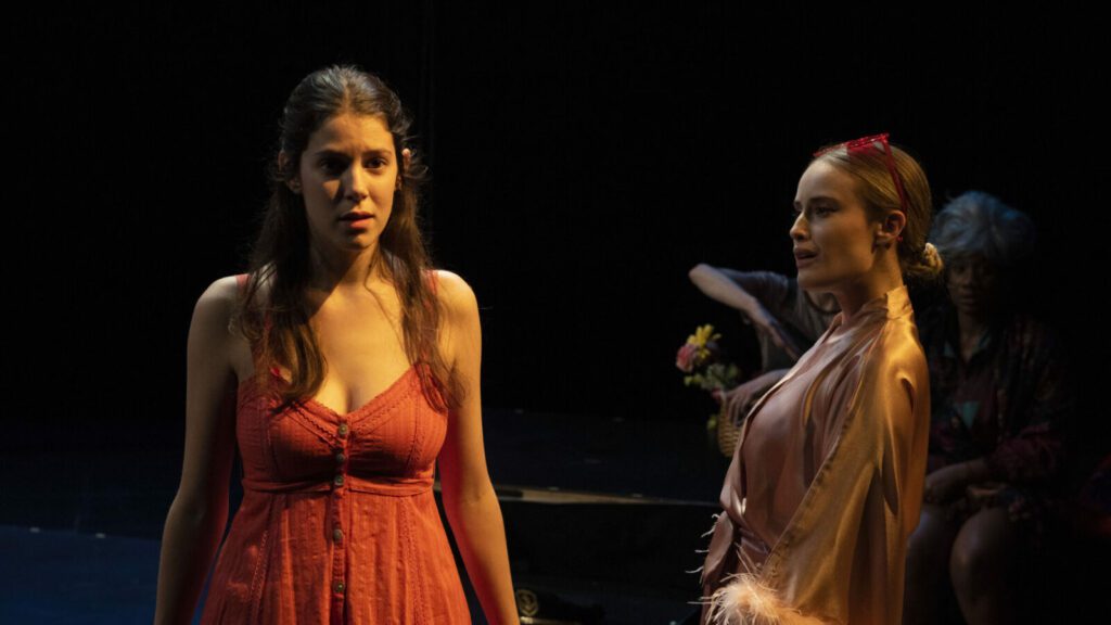 Actors on stage in a play, one in a red dress and another in a pink robe.