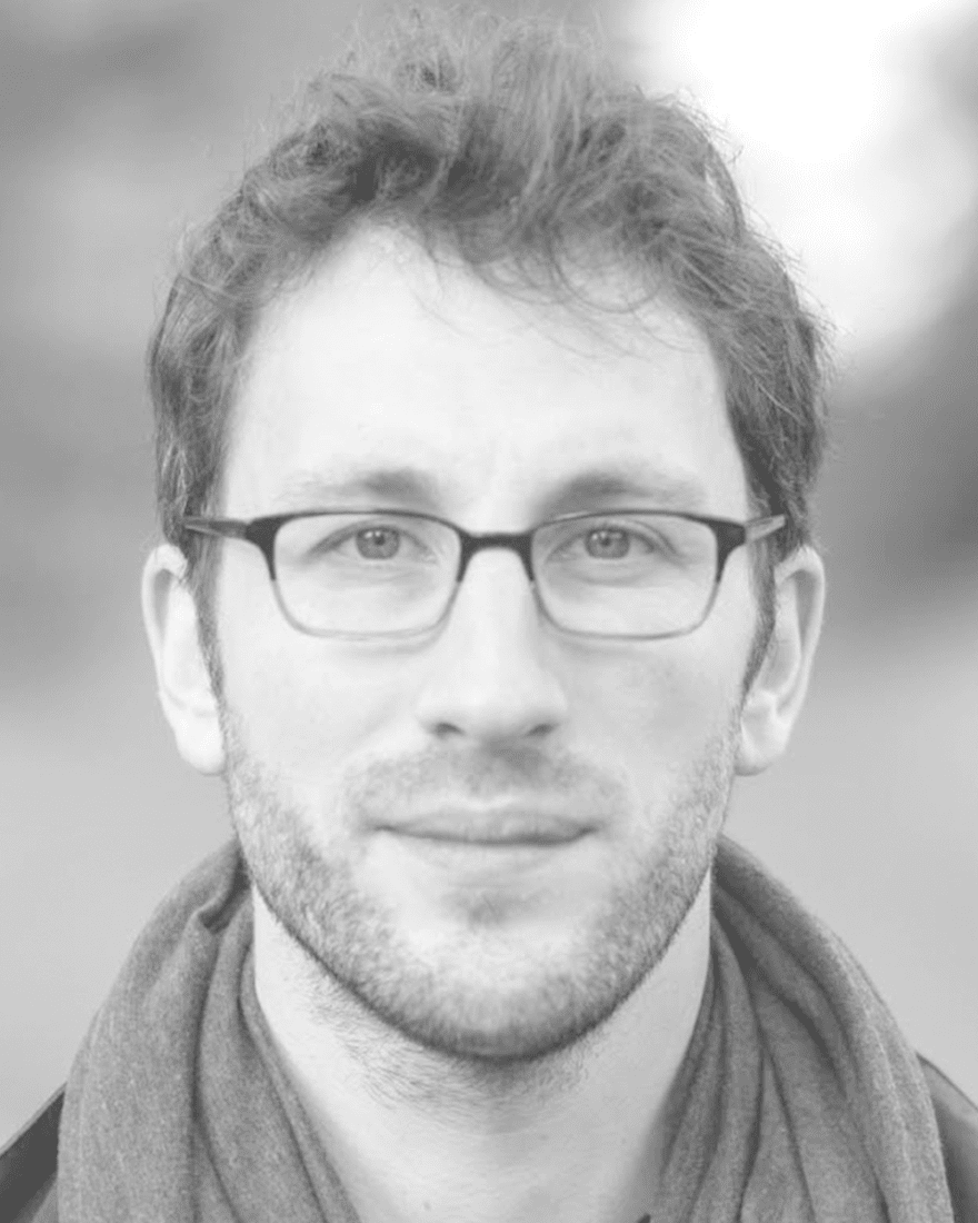 Headshot of Tom Costello wearing glasses and a scarf.