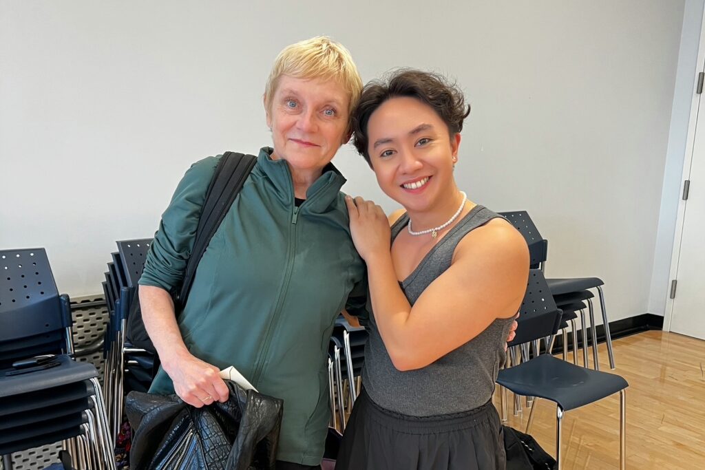 Two women pose in a dance studio; one in dance attire, the other in casual wear.