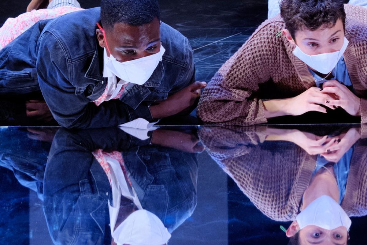 Two masked actors reflected on a glossy stage during a production.