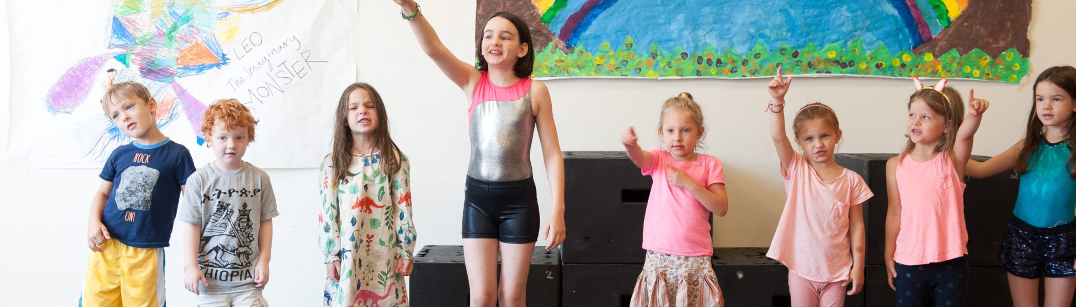 Kids in a line, some pointing, in front of children's art: rainbow and "LEO The Imaginary MONSTER".