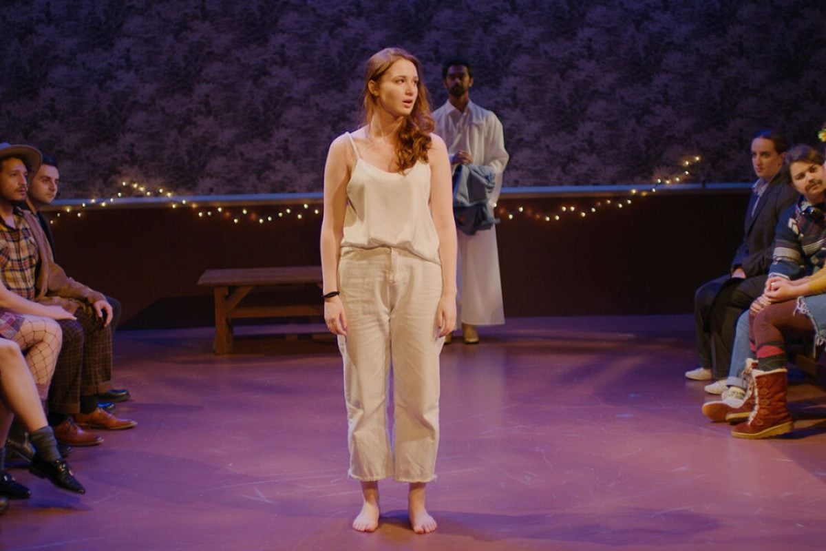 Play production scene: Woman stands center stage, surrounded by audience.