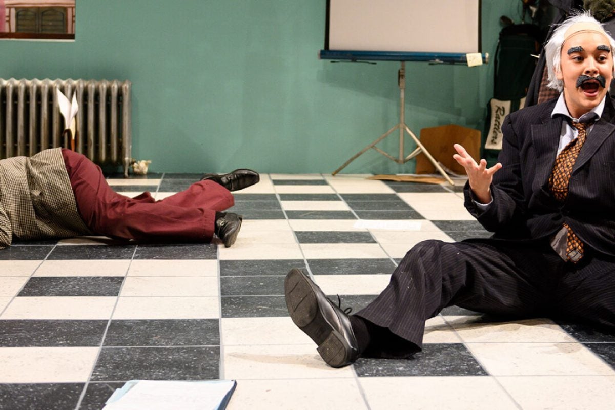 Play actors in costume sprawled on a checkered floor during a production.