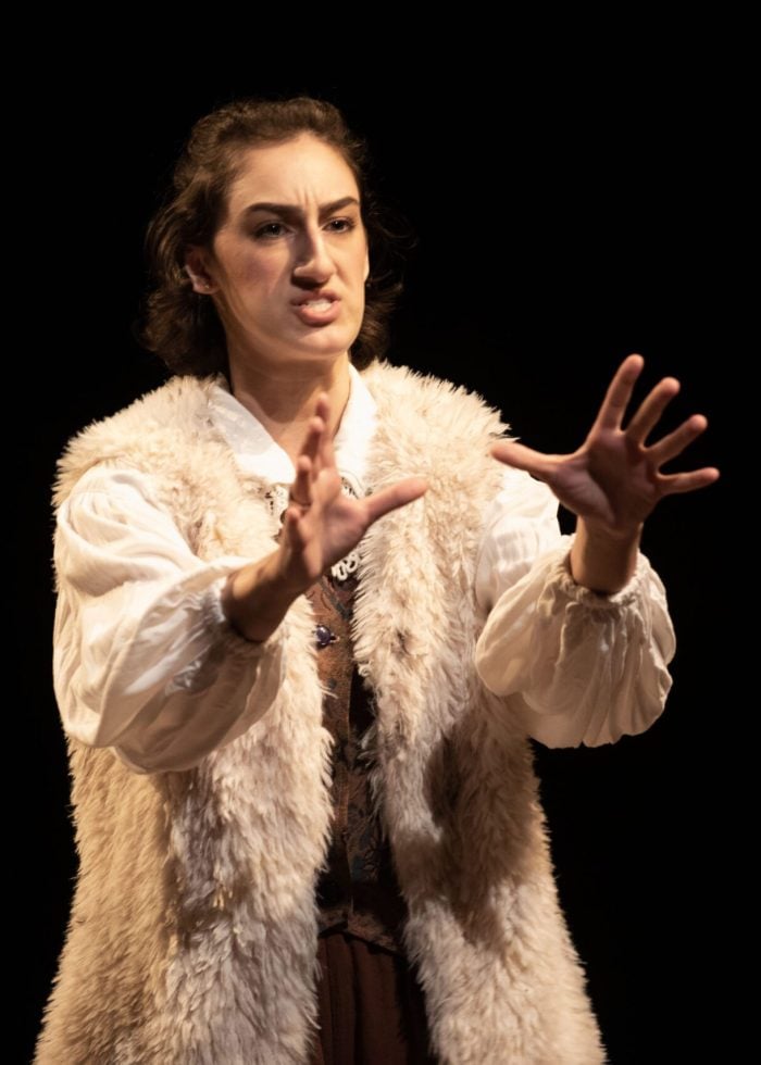 Actress performing in evening acting classes NYC, wearing a fur vest and white blouse, gesturing dramatically.