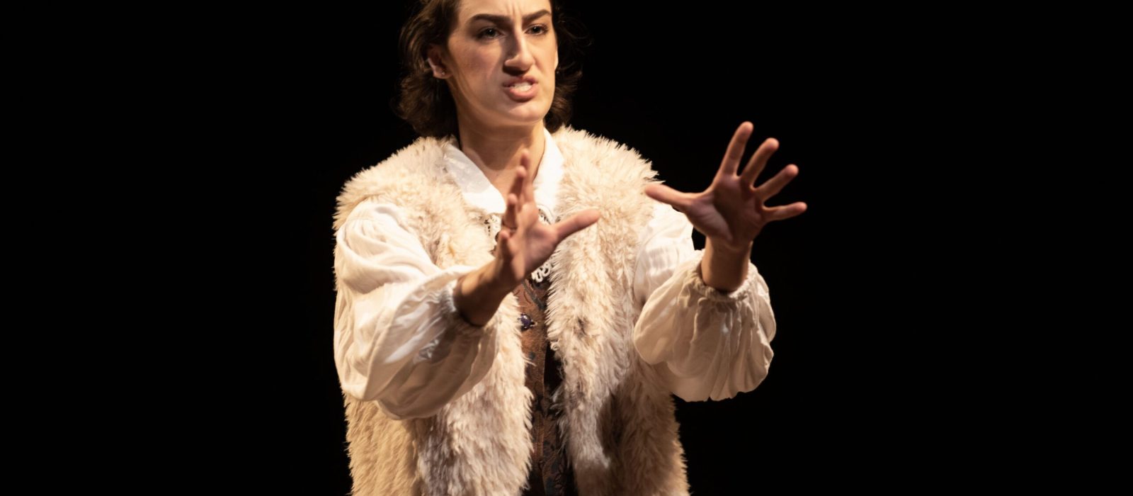 Actress performing in evening acting classes NYC, wearing a fur vest and white blouse, gesturing dramatically.