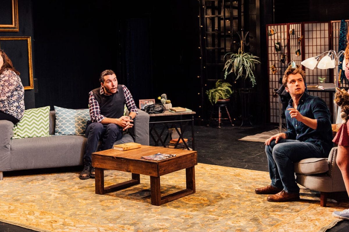 Theater production scene: Four actors on stage in a living room set, expressing surprise.