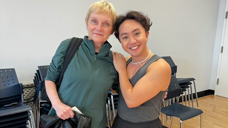 Two women pose in a dance studio; one in dance attire, the other in casual wear.