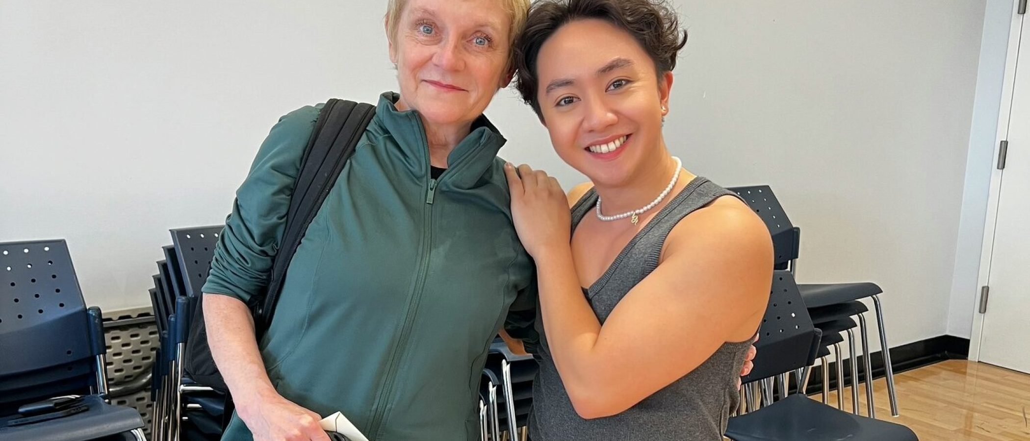 Two women pose in a dance studio; one in dance attire, the other in casual wear.