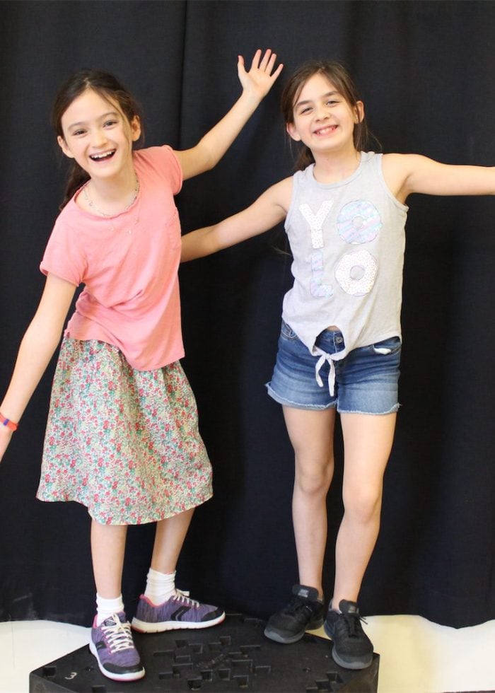 Two young girls pose with arms outstretched against a black backdrop. Sketch comedy fun!