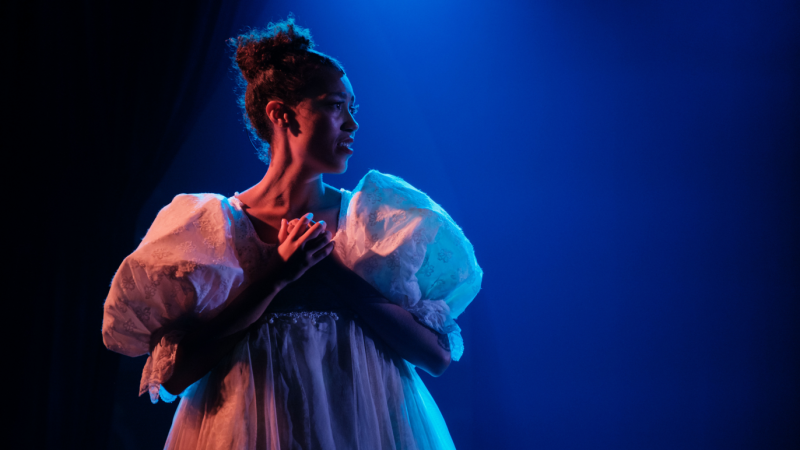 Performer in white dress on stage at the Atlantic Intensive, hands clasped.