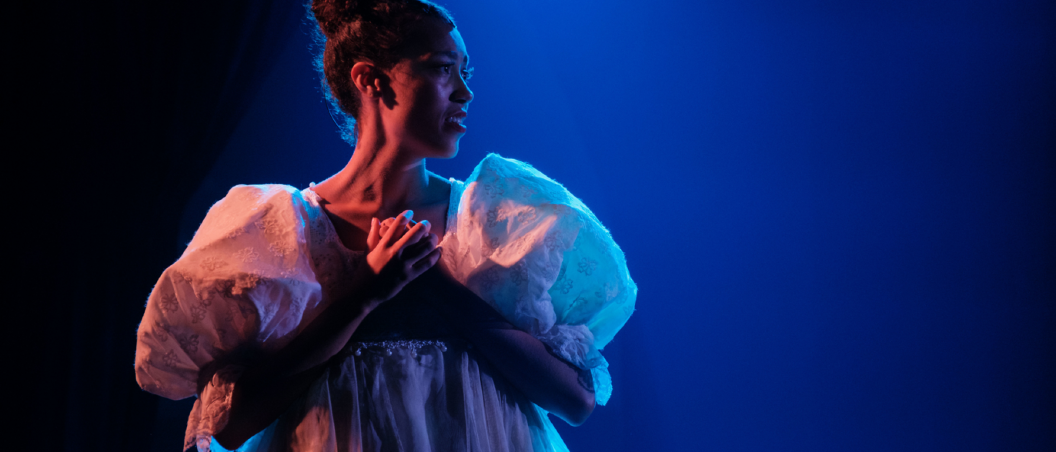 Performer in white dress on stage at the Atlantic Intensive, hands clasped.
