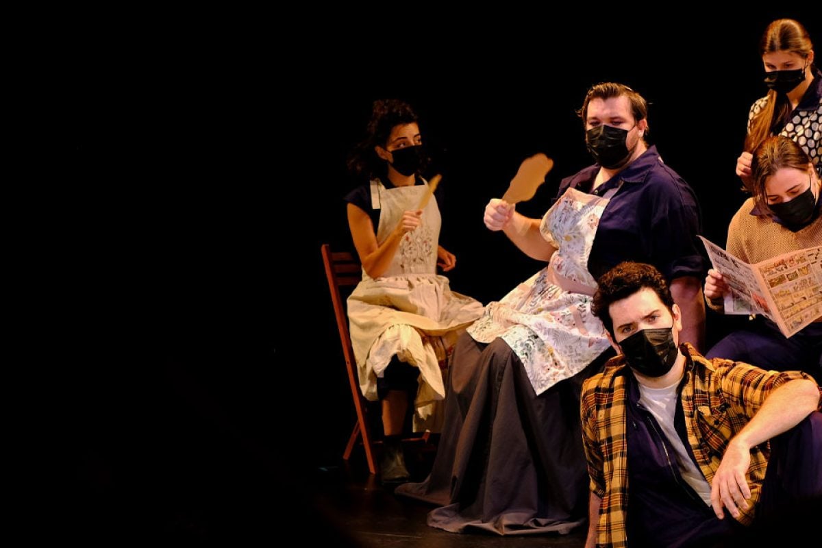 Theater production scene with masked actors in aprons and casual wear.