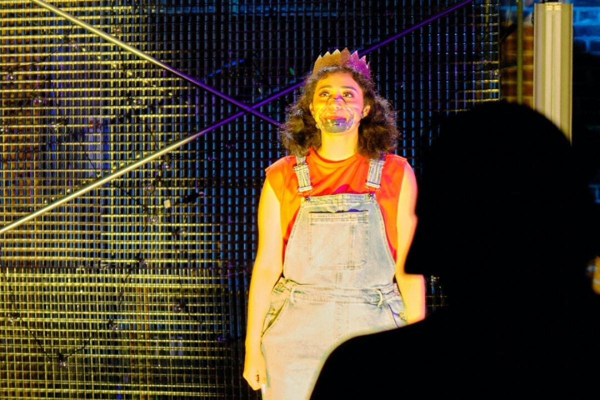 Woman in overalls and crown on stage during a theatrical production.